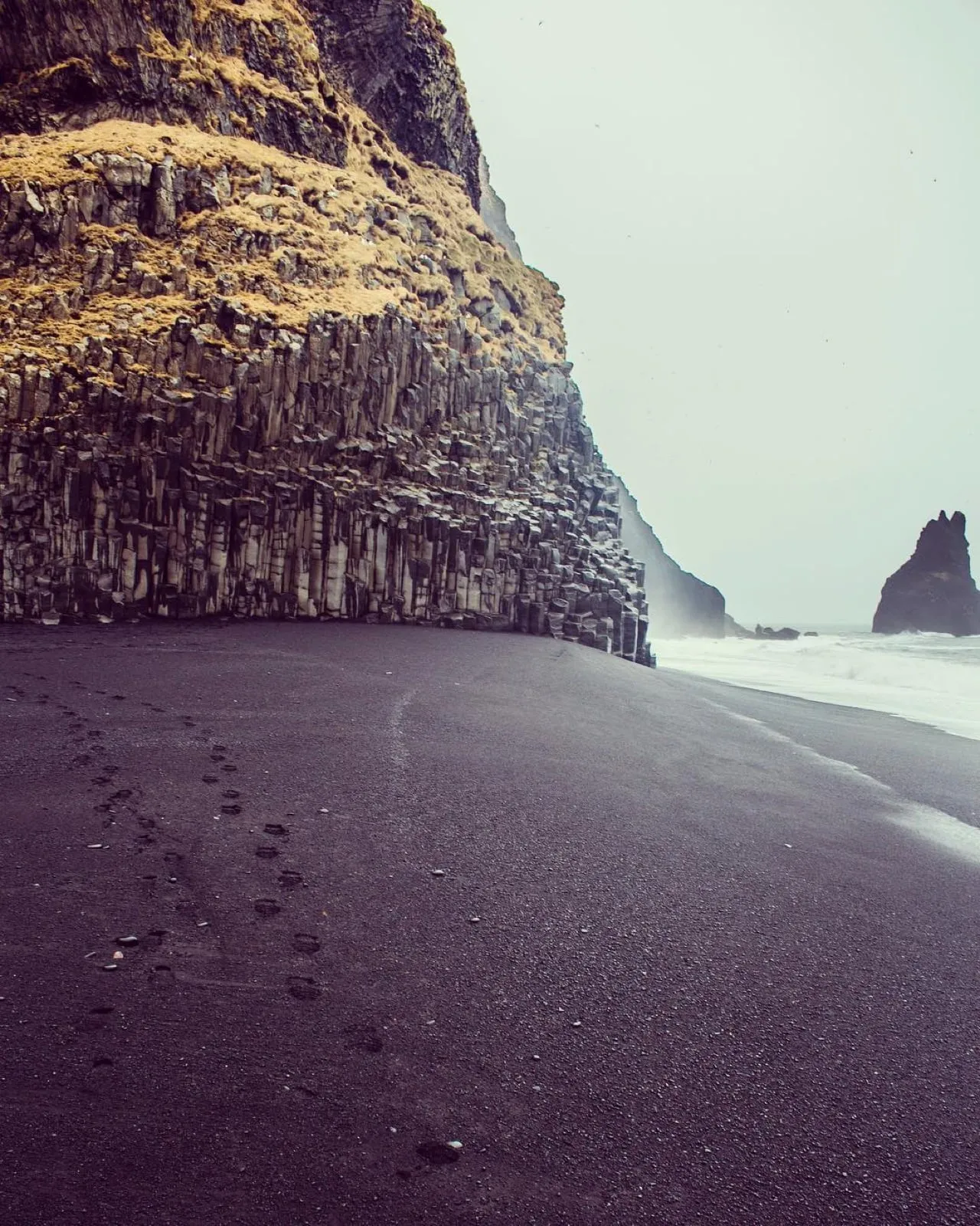Footprints on black sand leading toward tall basalt column cliffs and sea stacks near Vík