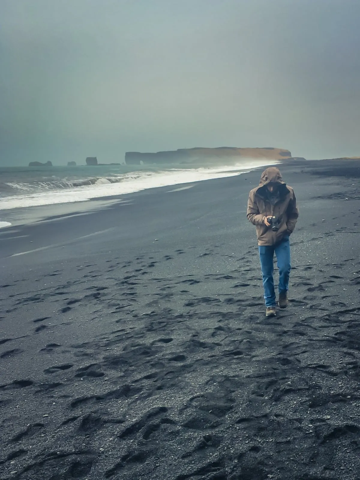 Michael walking toward the camera on a wide black sand beach near Vík, with rocky cliffs in the distance