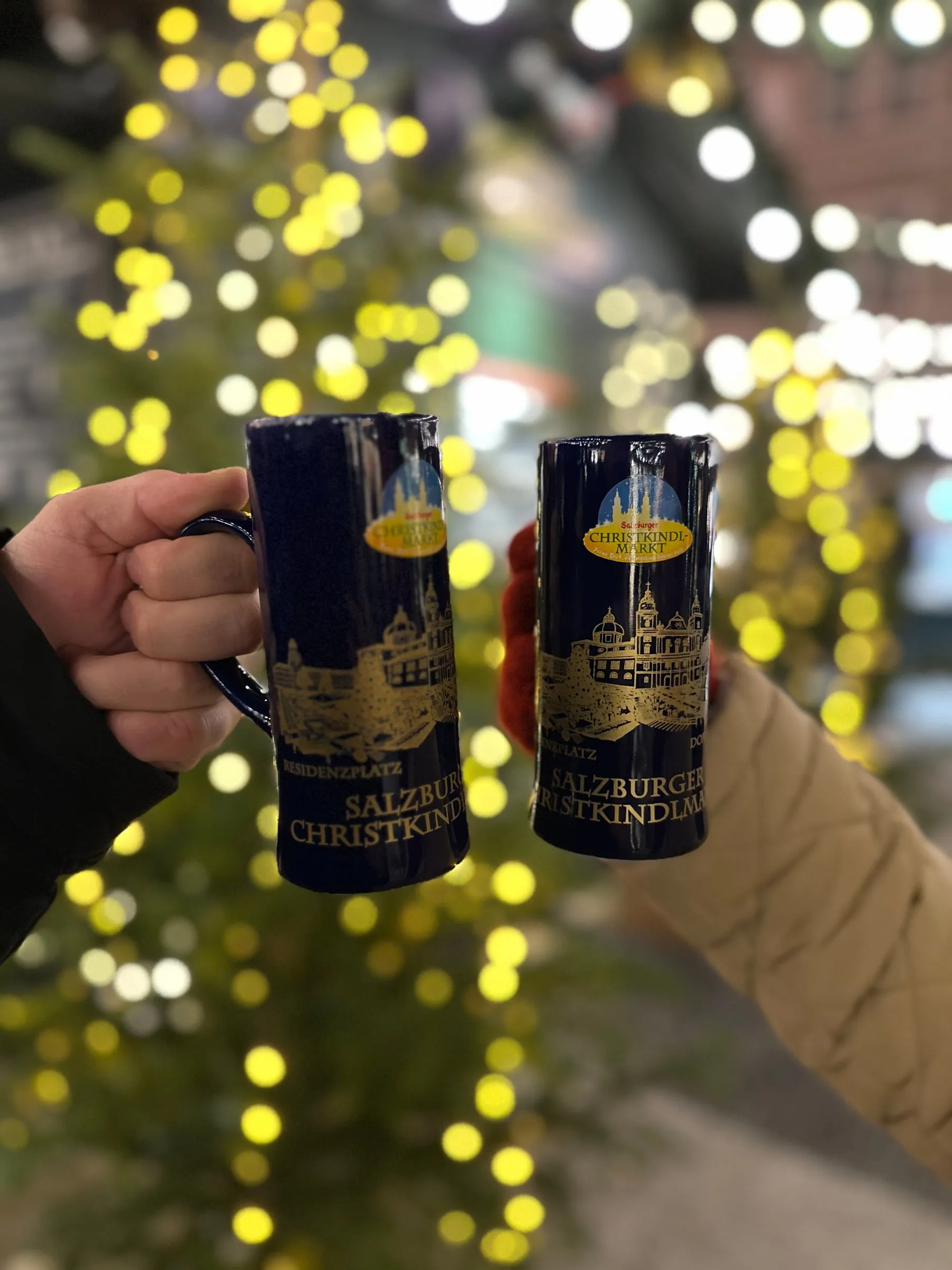 Two hands clinking blue Salzburg Christkindlmarkt glühwein mugs in front of a lit Christmas tree