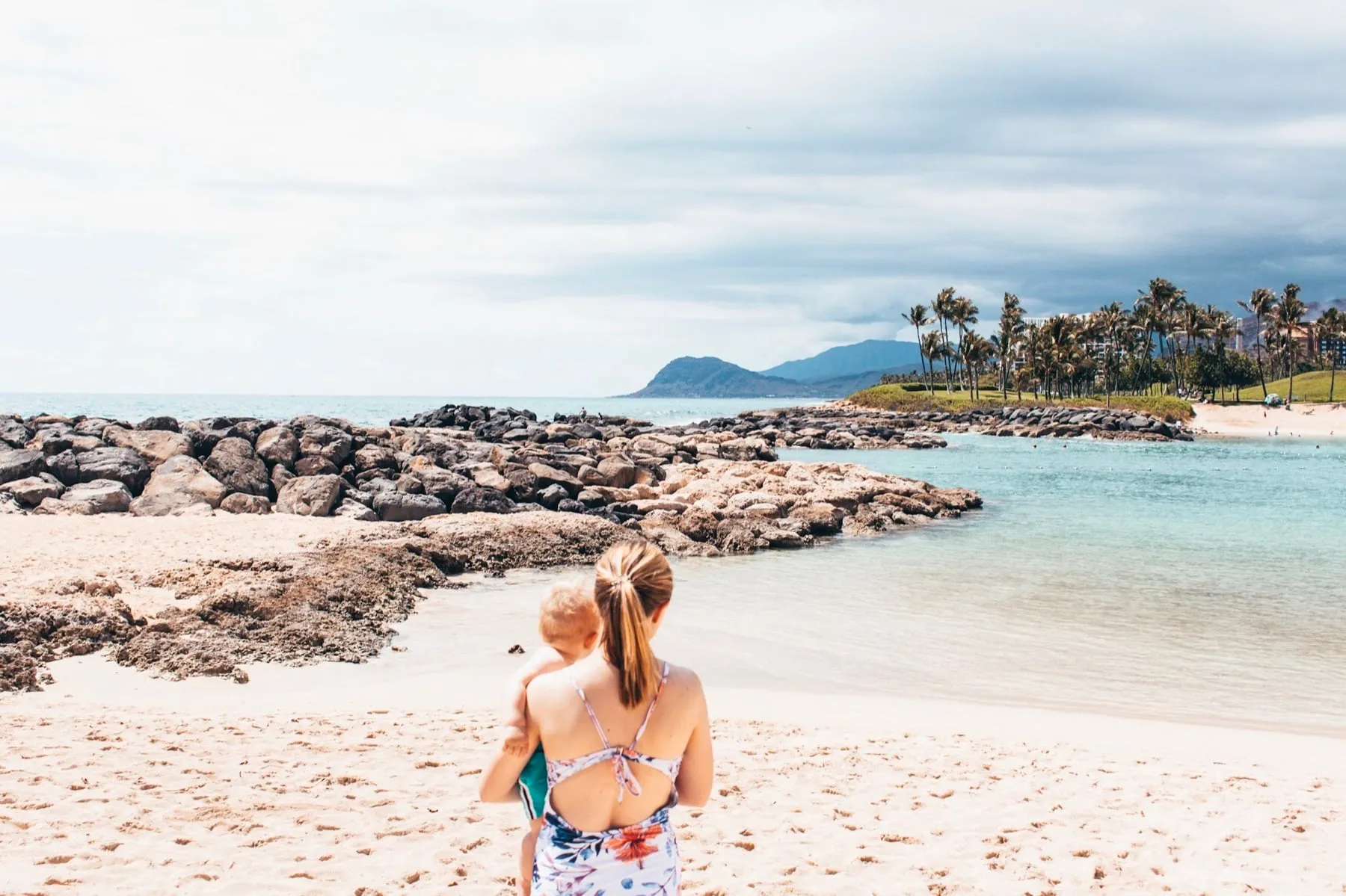 Mom holding our baby at the edge of a calm, clear lagoon on Oahu with palm trees in the distance