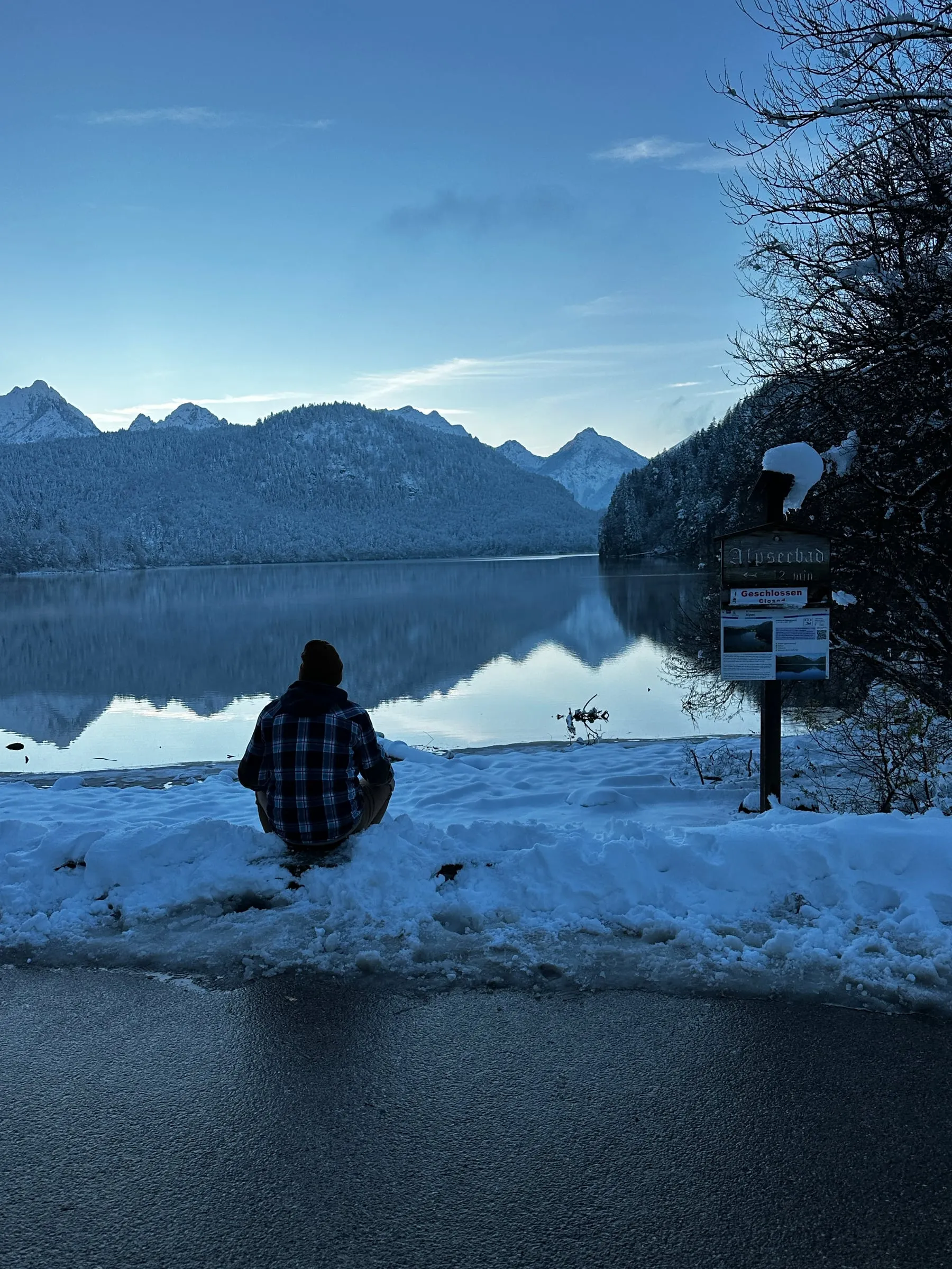 The calm Alpsee reflecting snowy mountains, with someone standing on the snowy shore