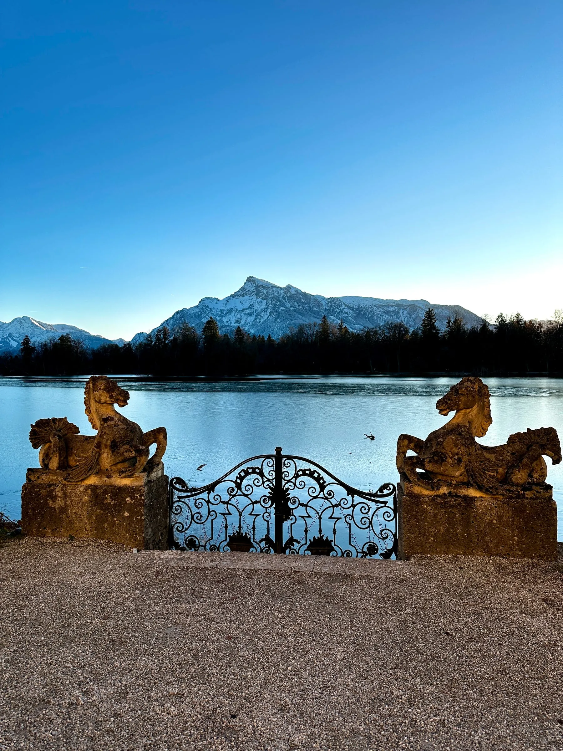 The lakeside gate of Schloss Leopoldskron, with two horse statues and snowy mountains behind the lake at sunset