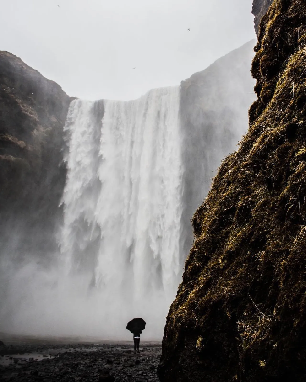 A tiny figure with an umbrella dwarfed by the misty curtain of Skógafoss