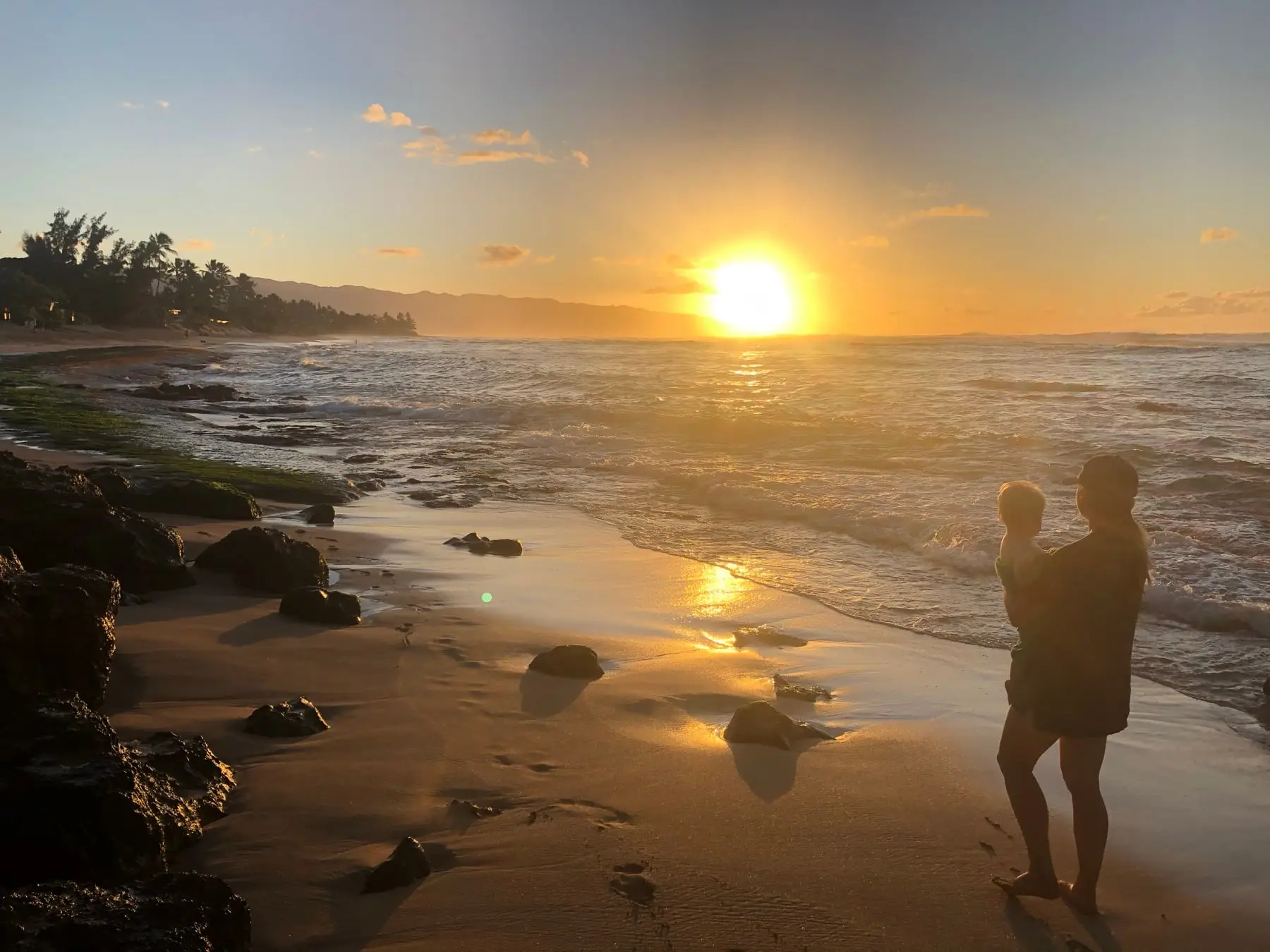 A parent holding a little one, silhouetted against a golden sunset on a Hawaii beach