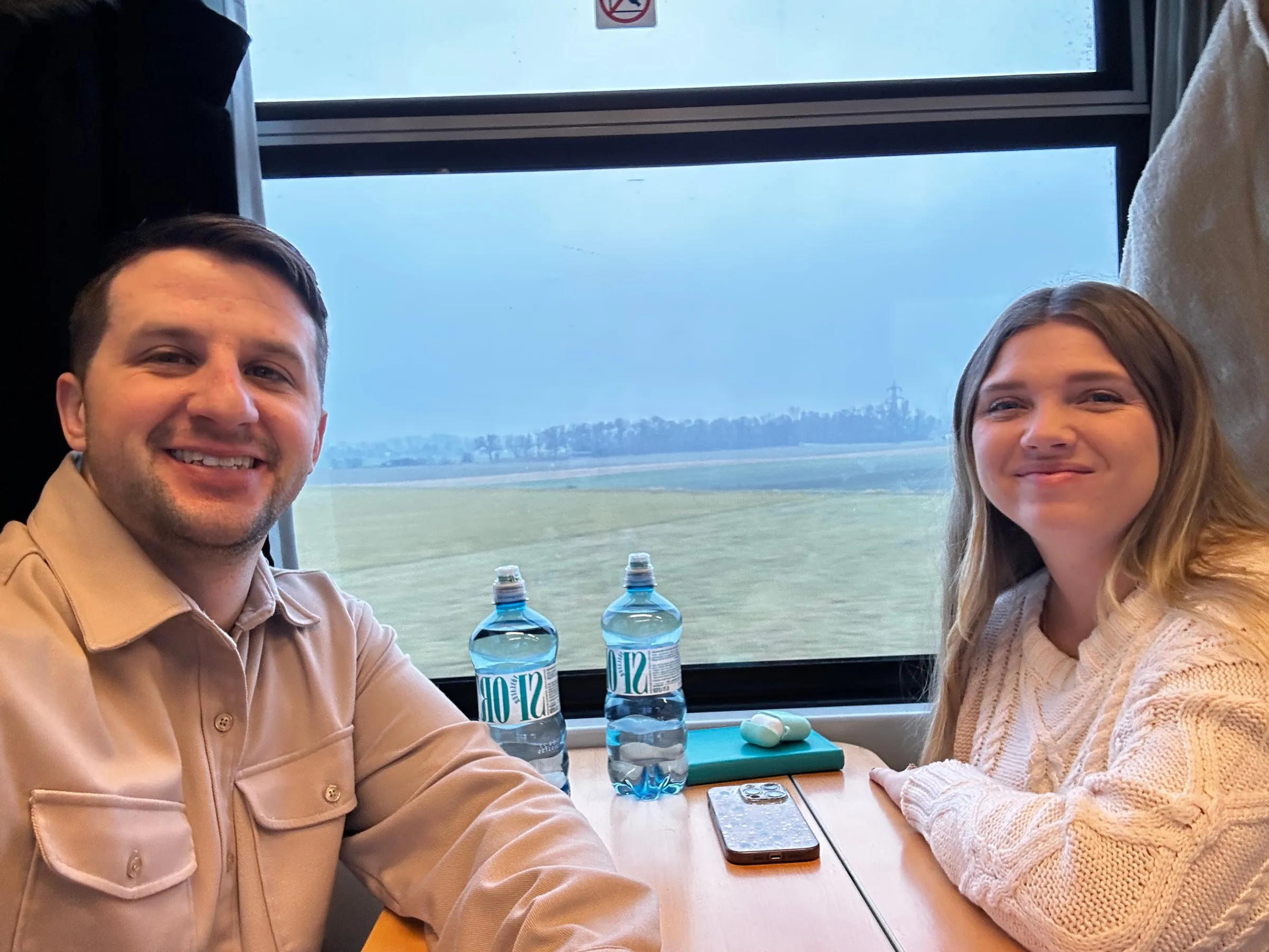 Michael and Chantelle at a table seat on a European train with water bottles and a window view of the countryside